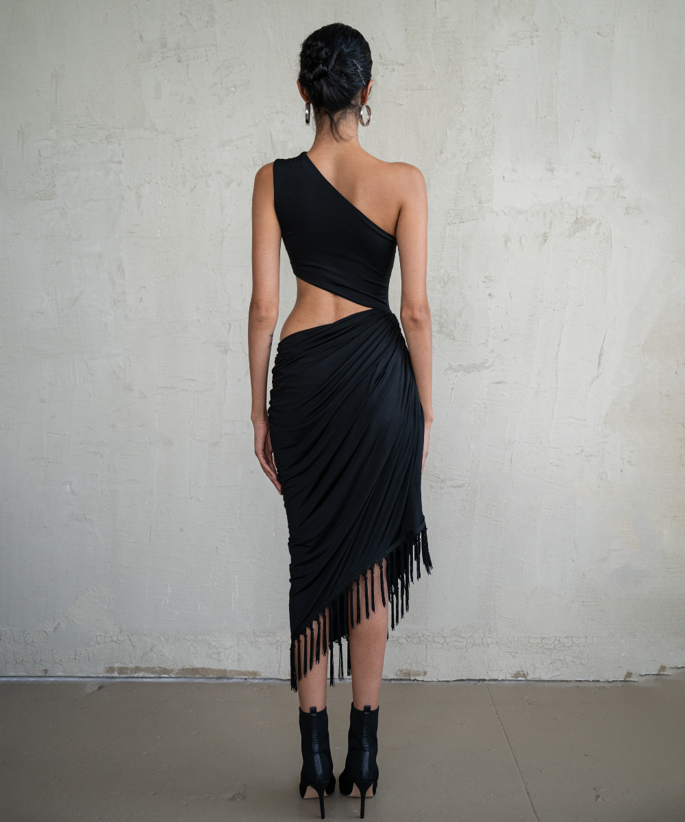 Woman wearing a black one-shoulder dress with fringe details against a plain background