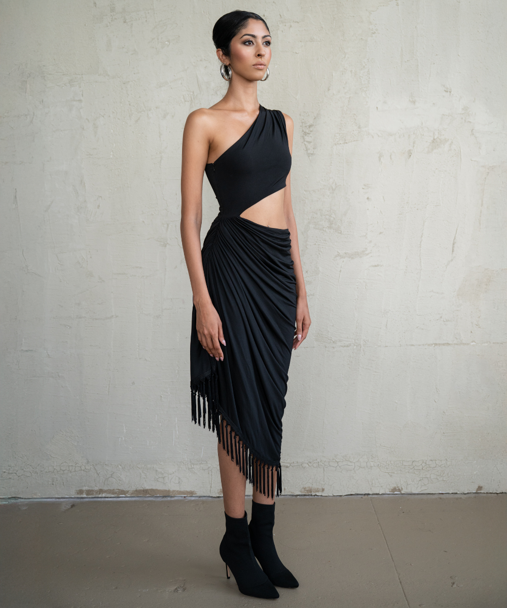 Woman wearing a black one-shoulder dress with fringe details against a plain background