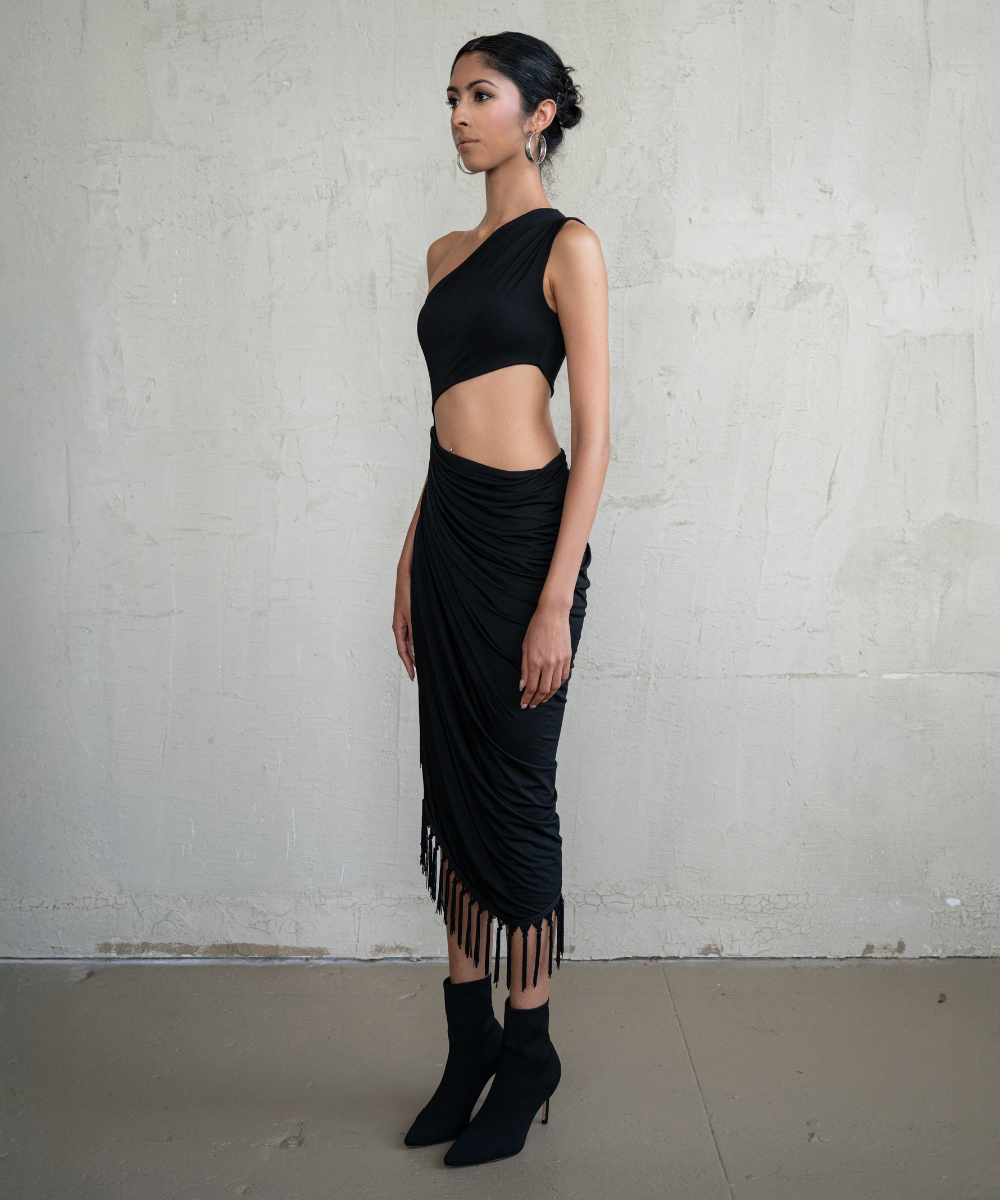 Woman wearing a black one-shoulder top and fringed skirt against a plain background