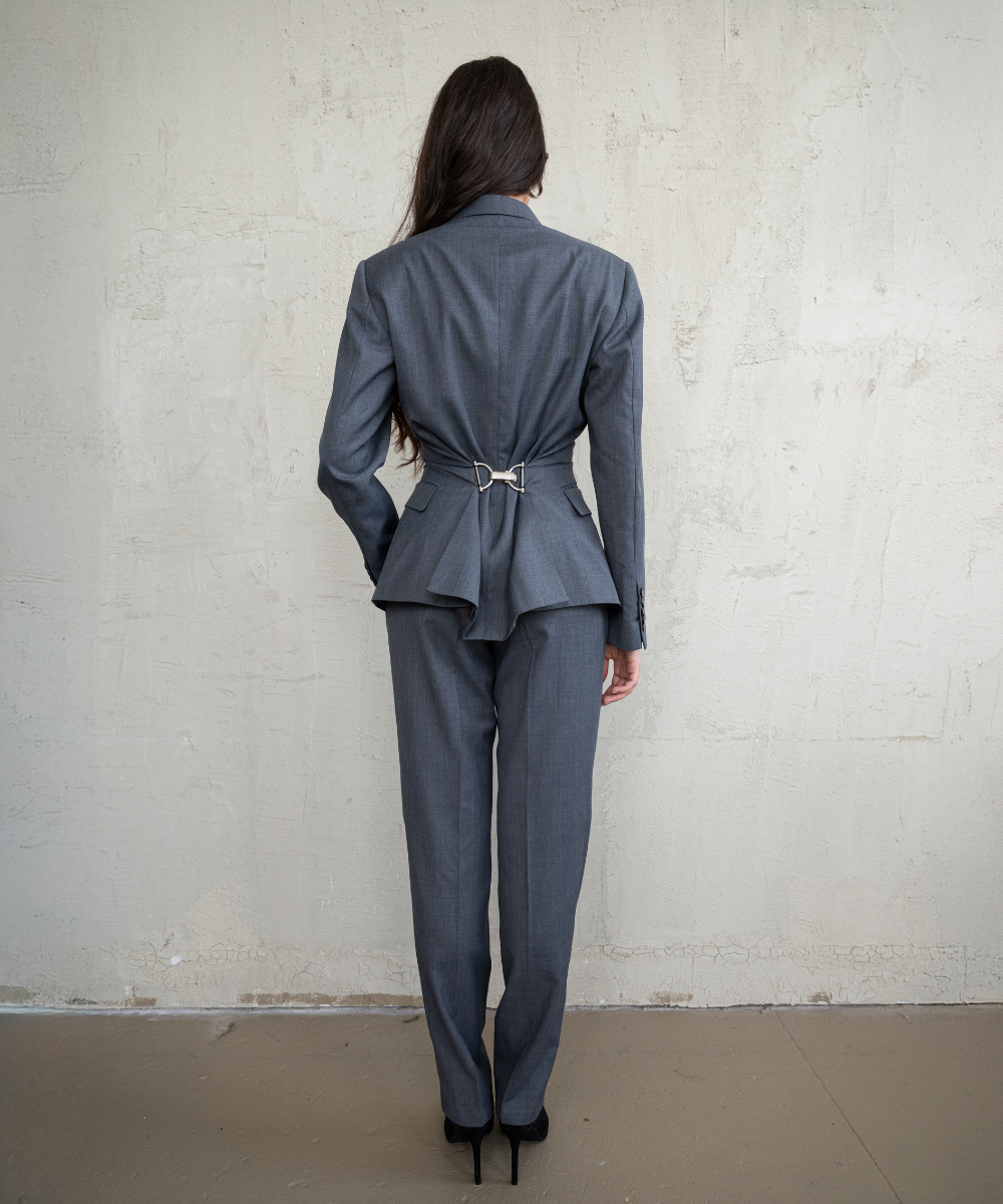 Woman wearing a gray suit standing against a white brick wall.